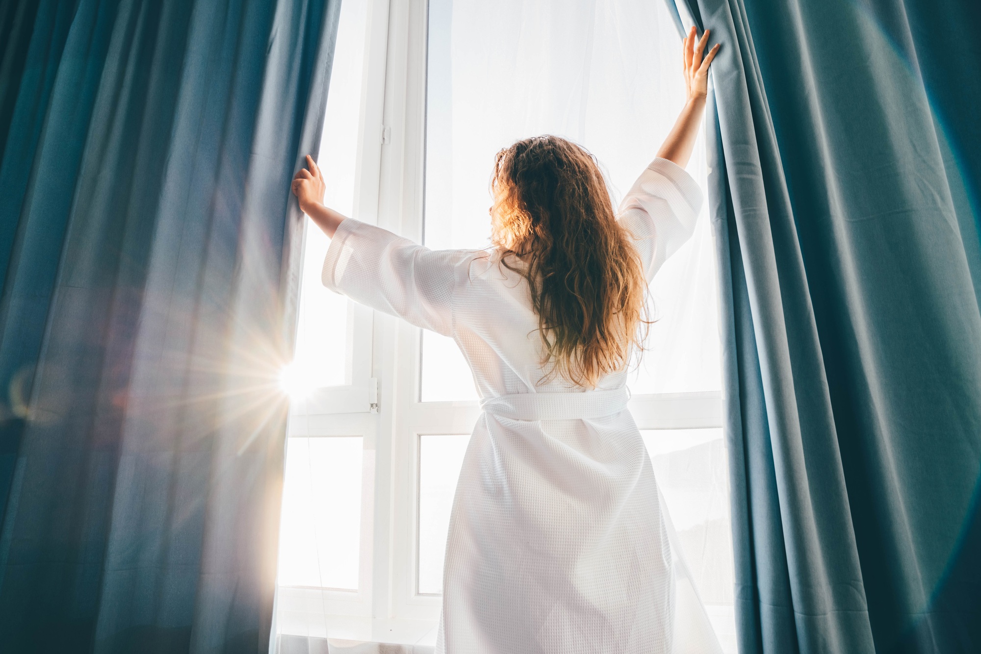 Woman in a bathrobe opening curtains in room at sunrise.